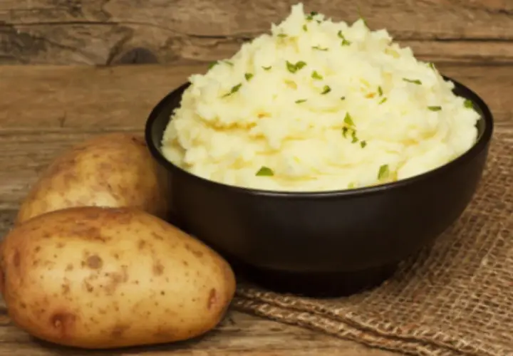 Un cremoso puré de patatas Thermomix, adornado con perejil picado, junto a patatas frescas sobre una mesa de madera.