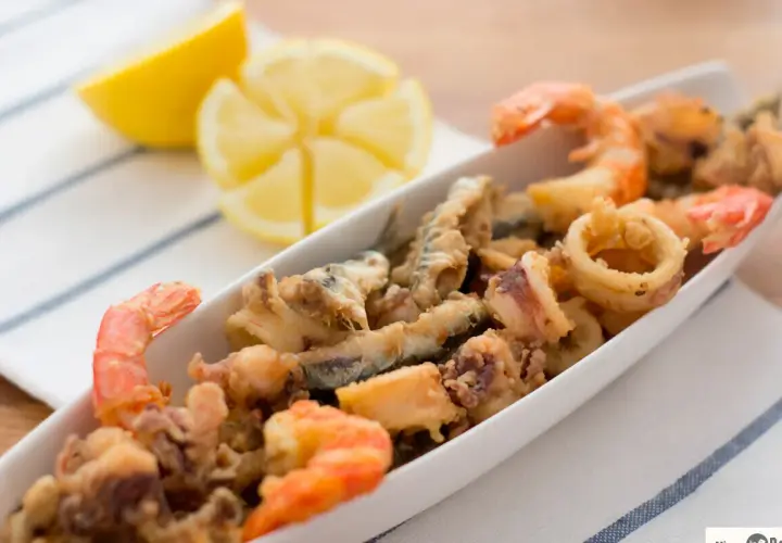 Deliciosa fritura de pescado con gambas y calamares, servida en una bandeja blanca con gajos de limón.