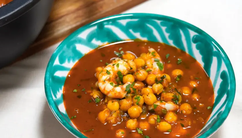 Un tazón de garbanzos con langostinos en una salsa rojiza, adornado con perejil picado. Un plato reconfortante y lleno de sabor.