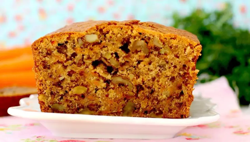Un delicioso bizcocho de zanahoria, con trozos de nueces y un color dorado apetitoso, servido en un plato blanco sobre un mantel floral.