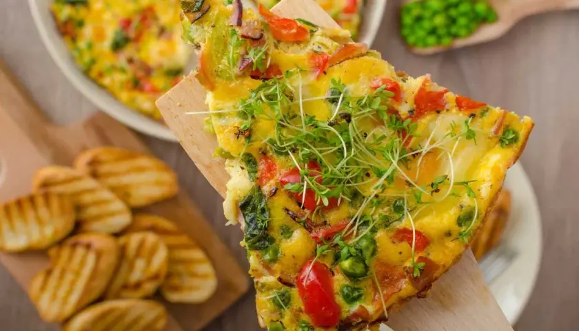 Rebanada de frittata de verduras con guisantes, pimientos y brotes frescos, servida con patatas a la parrilla.