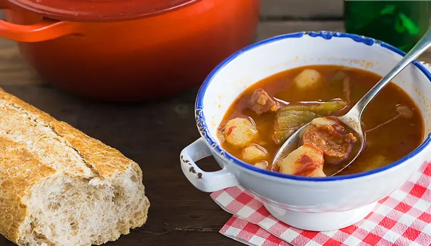 Un plato humeante de patatas a la riojana, con chorizo y pimientos, acompañado de pan rústico y una olla roja.