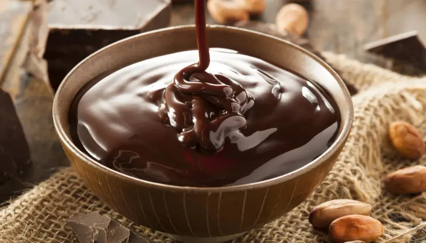Un chorro de ganache de chocolate cae en un cuenco, rodeado de trozos de chocolate y granos de cacao.