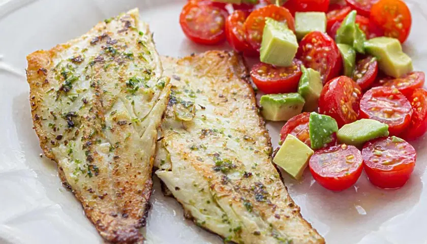 Dos filetes de lubina a la plancha con hierbas, acompañados de una ensalada fresca de tomates cherry y aguacate en cubos.