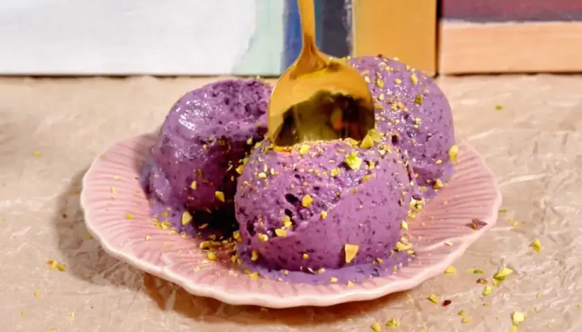 Tres bolas de helado casero de arándanos con pistachos picados, servidas en un plato rosa. Una cuchara dorada se hunde en el helado.