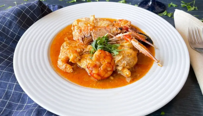 Un plato de rap a la marinera con gambas y cigalas en una salsa rojiza, decorado con perejil fresco.