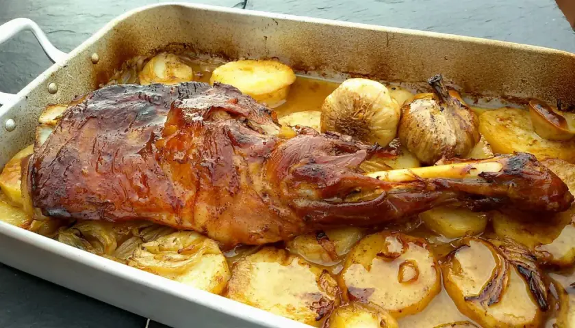 Paletilla de cordero asada con patatas y ajos dorados en una bandeja. Jugosa y apetitosa, lista para servir.