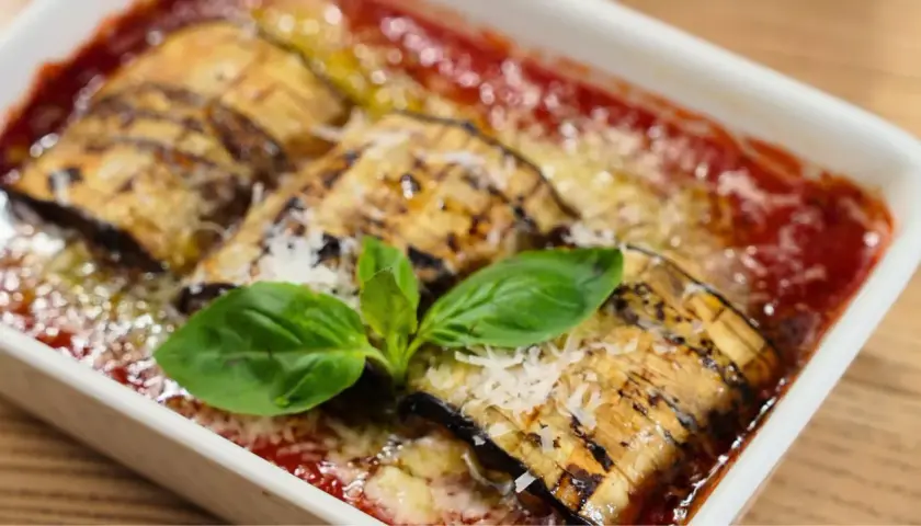 Delicioso pastel de berenjenas gratinado con queso y bañado en salsa de tomate, adornado con hojas de albahaca fresca.