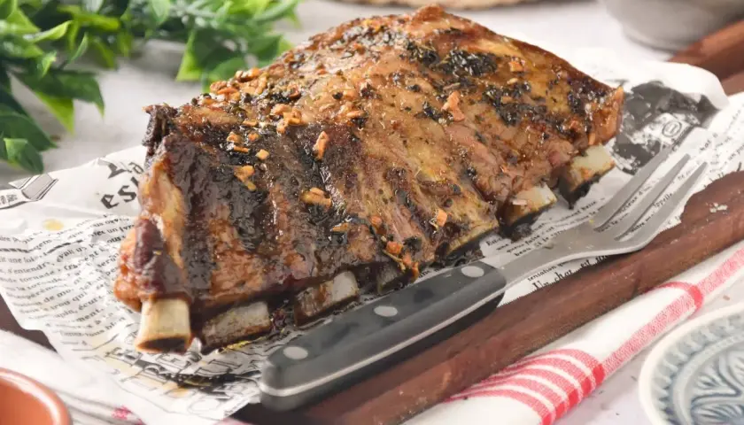 Costillas de churrasco de cerdo glaseadas, servidas sobre papel con texto y acompañadas de un tenedor y cuchillo.