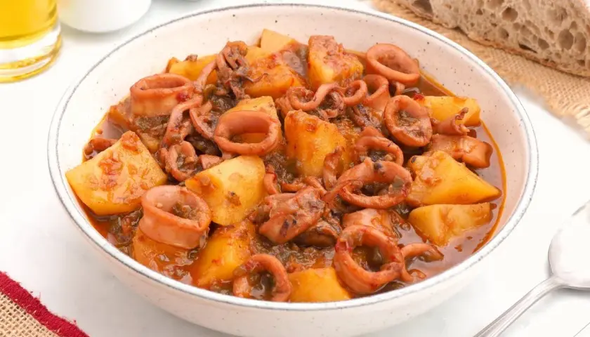 Un tazón de patatas con calamares en salsa, acompañado de pan rústico y un vaso de cerveza.