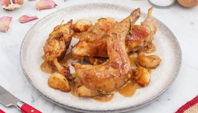 Delicioso conejo al ajillo servido en un plato, con ajos enteros asados y salsa dorada.