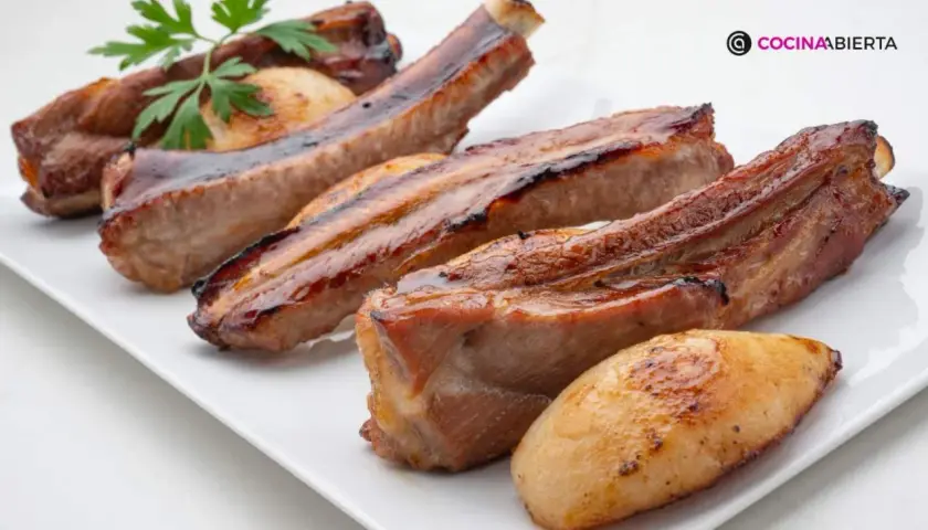 Costillas de cerdo jugosas y doradas, servidas en una bandeja blanca con patatas asadas y una ramita de perejil.