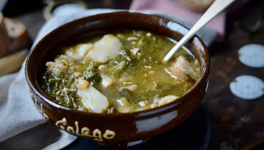 Un cuenco de barro con la palabra "Gallego" escrita, lleno de un reconfortante caldo gallego con patatas, grelos y trozos de pan.