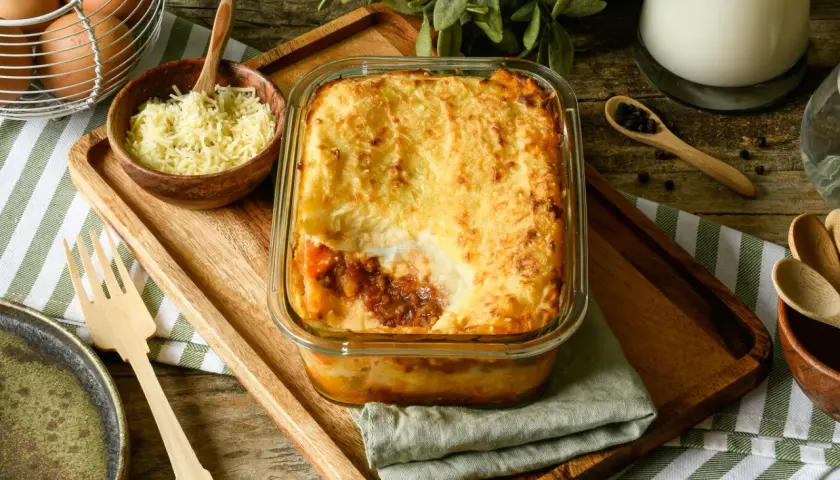 Pastel de carne picada recién horneado, con una capa dorada de puré de patatas y queso gratinado. Se sirve en una fuente de cristal sobre una tabla de madera.