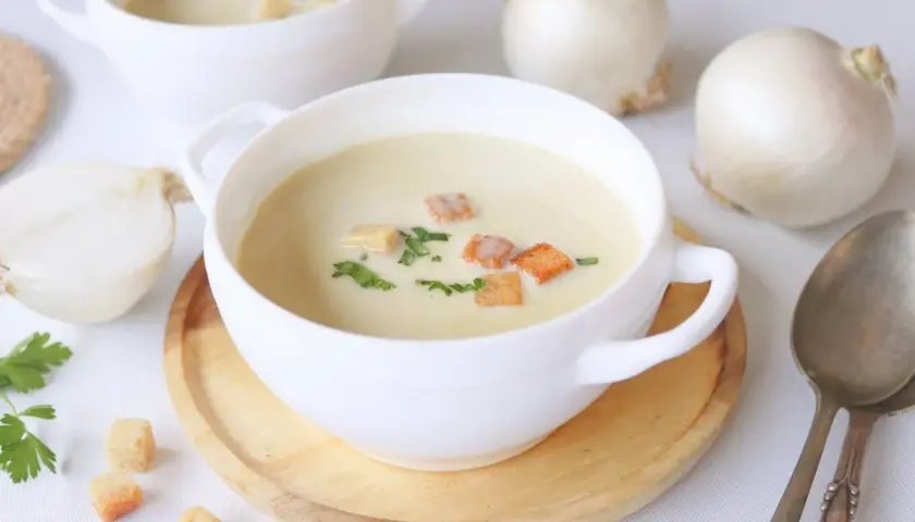 Crema de cebolla cremosa, adornada con picatostes dorados y perejil fresco. Cebollas blancas enteras y cortadas a medias rodean los cuencos.