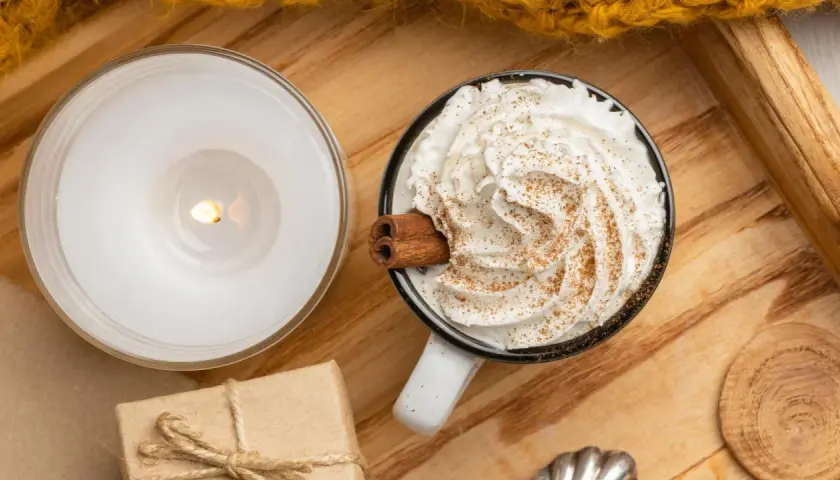 Taza de bebida caliente con abundante **montar nata**, canela y una rama de canela. Una vela encendida y un regalo completan la escena acogedora.