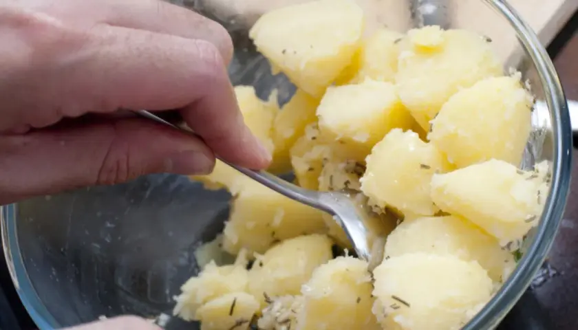 Manos mezclando patatas cocidas con hierbas en un bol de cristal.