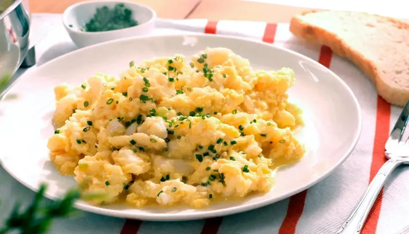 Revuelto de bacalao cremoso, espolvoreado con cebollino fresco, servido en un plato blanco con una rebanada de pan tostado al lado.