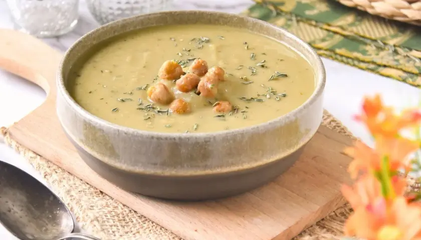 Un tazón de cremosa crema de garbanzos, decorada con garbanzos enteros y hierbas, servida en una tabla de madera.