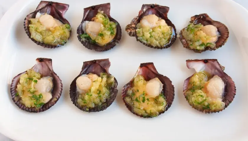 Ocho zamburiñas al horno, listas para disfrutar, con un relleno dorado y perejil picado.
