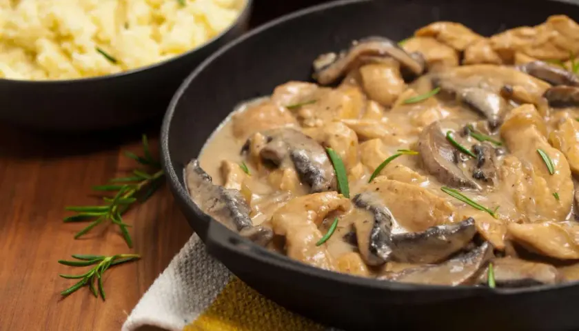 Delicioso pollo con champiñones en salsa cremosa, servido en sartén de hierro fundido, con puré de patatas al lado.