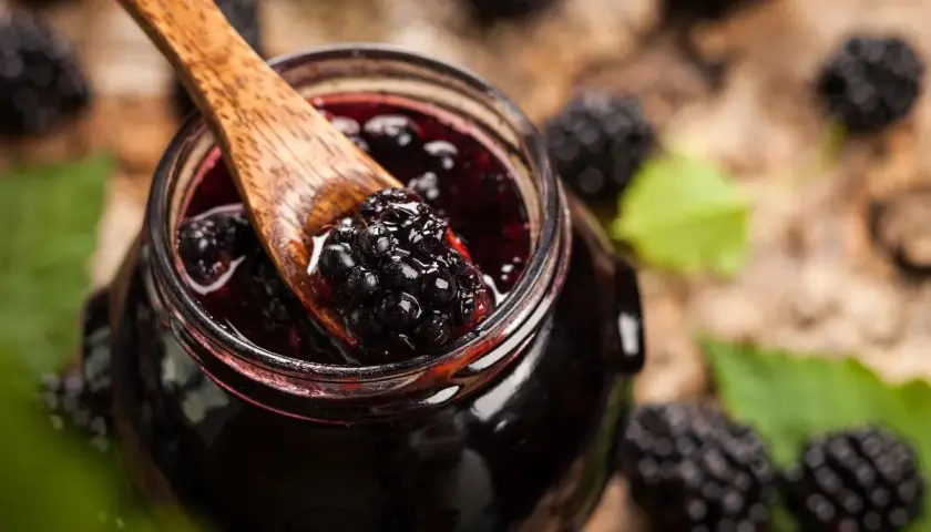 Cuchara de madera recoge mermelada de moras silvestres de un tarro, rodeado de moras frescas y hojas verdes.