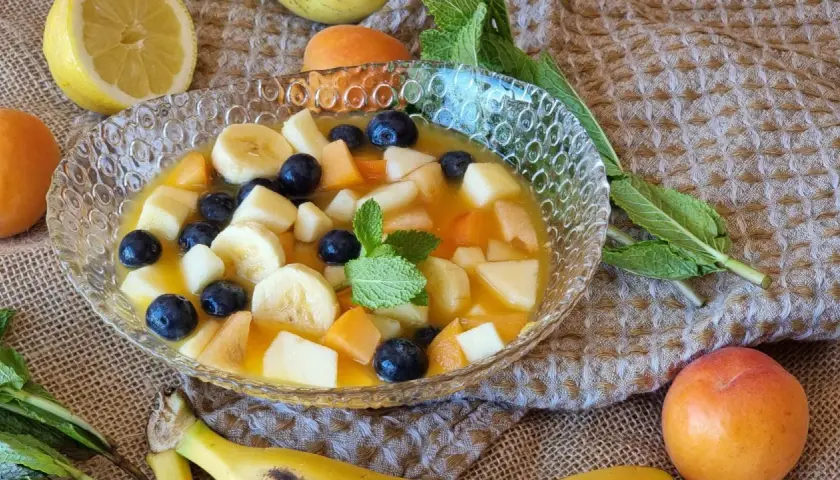 Deliciosa macedonia de frutas con plátano, arándanos, melocotón y manzana en un cuenco de cristal.