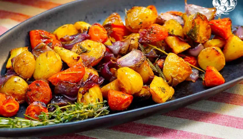 Un plato de verduras al horno, con patatas, zanahorias y cebolla morada, adornado con romero fresco.