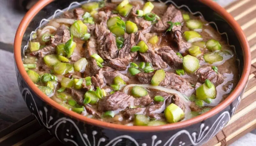 Un reconfortante plato de ternera guisada con trozos de carne tierna, fideos y espárragos verdes, servido en un cuenco rústico.