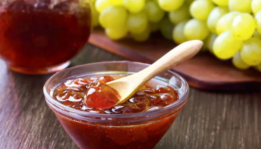 Un cuenco de cristal con mermelada de uvas casera y una cuchara de madera, junto a un racimo de uvas verdes.