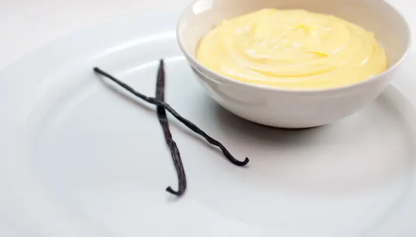 Crema inglesa cremosa en un tazón, con dos vainas de vainilla oscuras al lado.