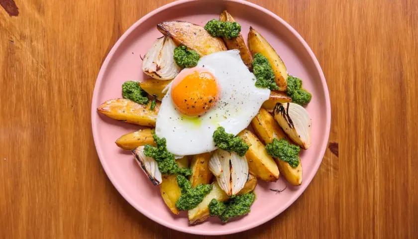 Patatas al horno doradas con huevo frito, cebolla asada y pesto verde, servidas en un plato rosa sobre una mesa de madera.