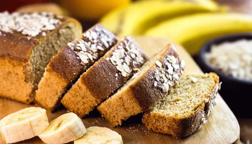 Rebanadas de bizcocho de plátano sin azúcar, cubiertas con avena, junto a plátanos frescos y un tazón de avena.