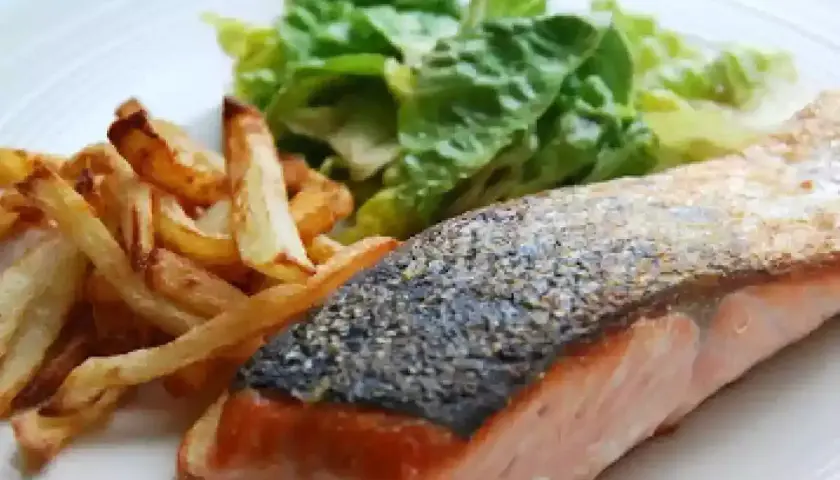 Un jugoso salmón a la plancha con piel crujiente, acompañado de patatas fritas doradas y una fresca ensalada verde.