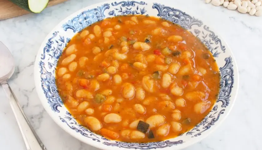Un plato de alubias blancas con verduras, cocinadas en una salsa sabrosa. Se ven trozos de pimiento, zanahoria y calabacín.