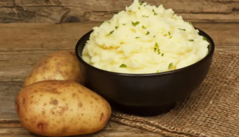 Un cremoso puré de patatas Thermomix, adornado con perejil picado, junto a patatas frescas sobre una mesa de madera.