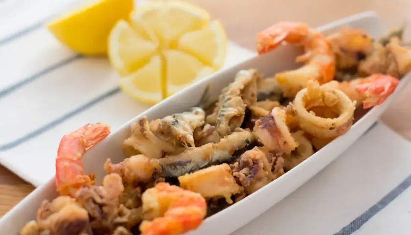 Deliciosa fritura de pescado con gambas y calamares, servida en una bandeja blanca con gajos de limón.