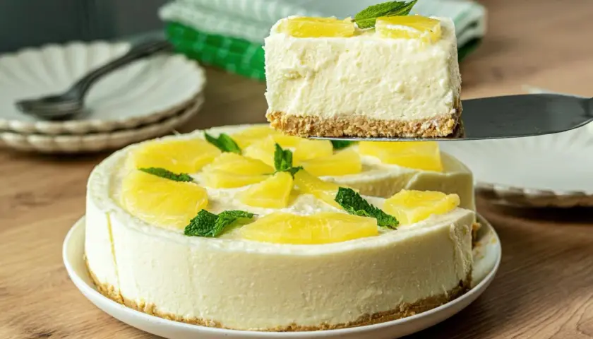 Una porción de tarta de piña sin horno, cremosa y decorada con piña y menta, se levanta de la tarta completa.