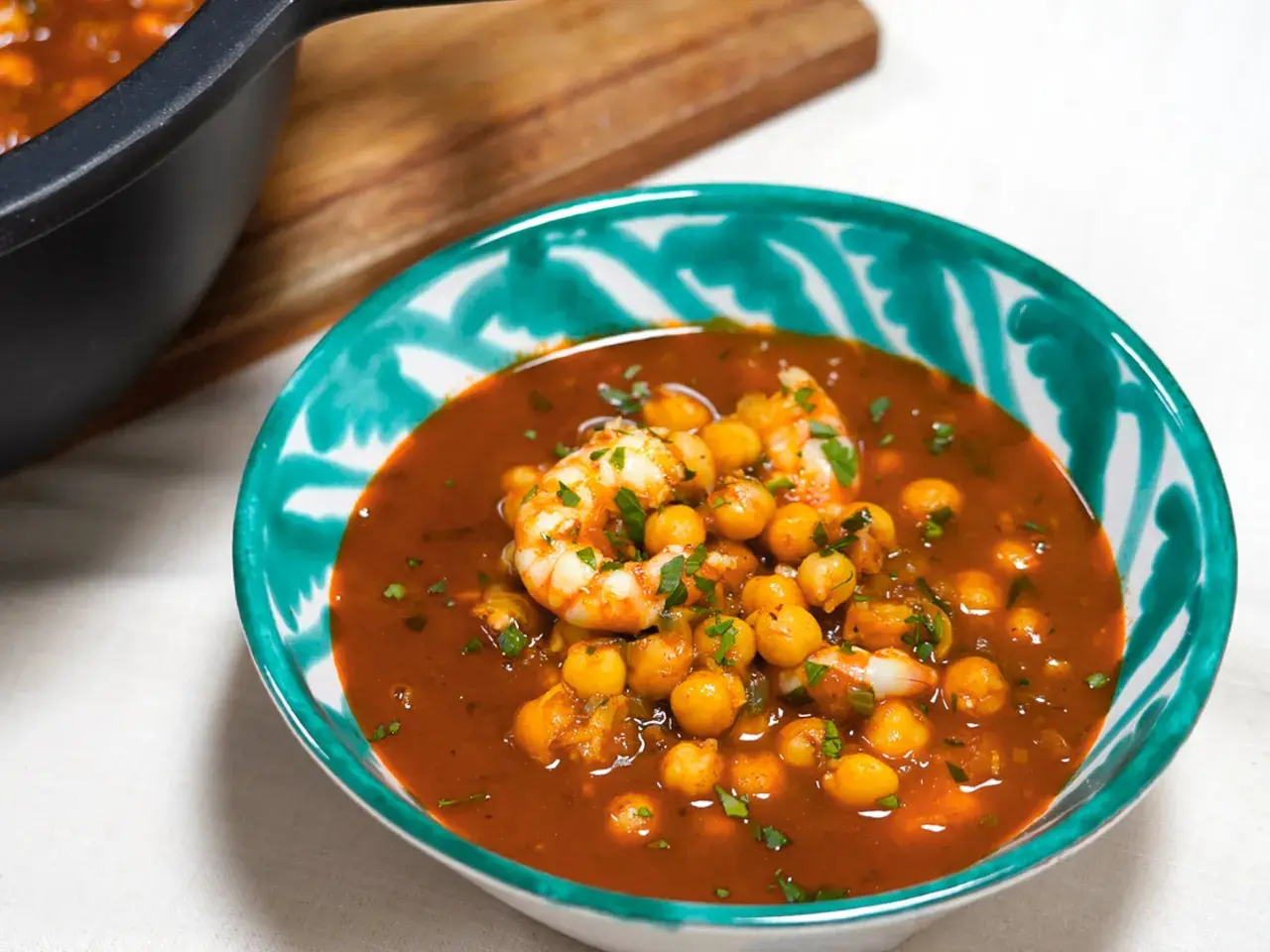 Un tazón de garbanzos con langostinos en una salsa rojiza, adornado con perejil picado. Un plato reconfortante y lleno de sabor.