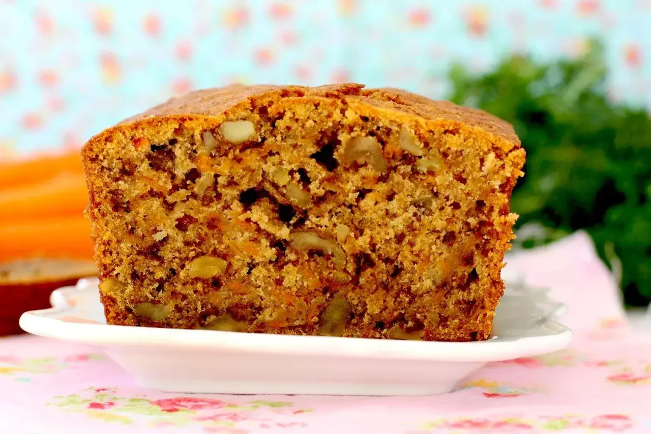 Un delicioso bizcocho de zanahoria, con trozos de nueces y un color dorado apetitoso, servido en un plato blanco sobre un mantel floral.