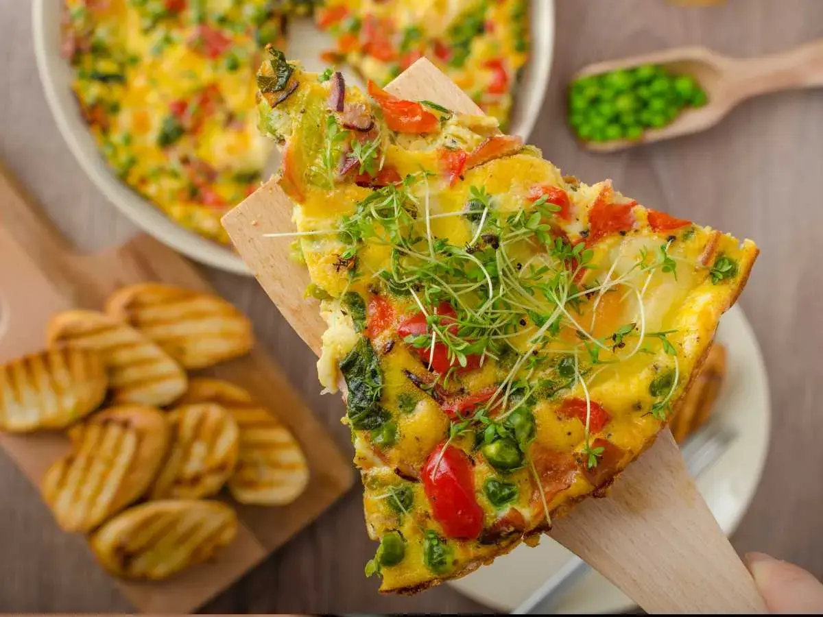 Rebanada de frittata de verduras con guisantes, pimientos y brotes frescos, servida con patatas a la parrilla.