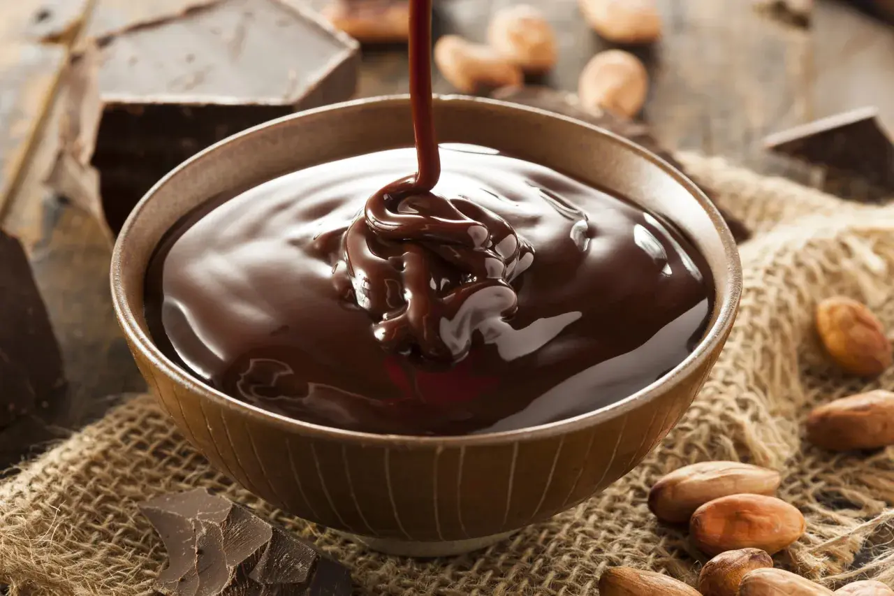 Un chorro de ganache de chocolate cae en un cuenco, rodeado de trozos de chocolate y granos de cacao.