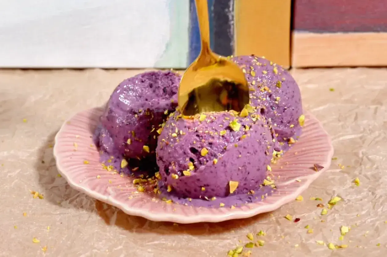 Tres bolas de helado casero de arándanos con pistachos picados, servidas en un plato rosa. Una cuchara dorada se hunde en el helado.