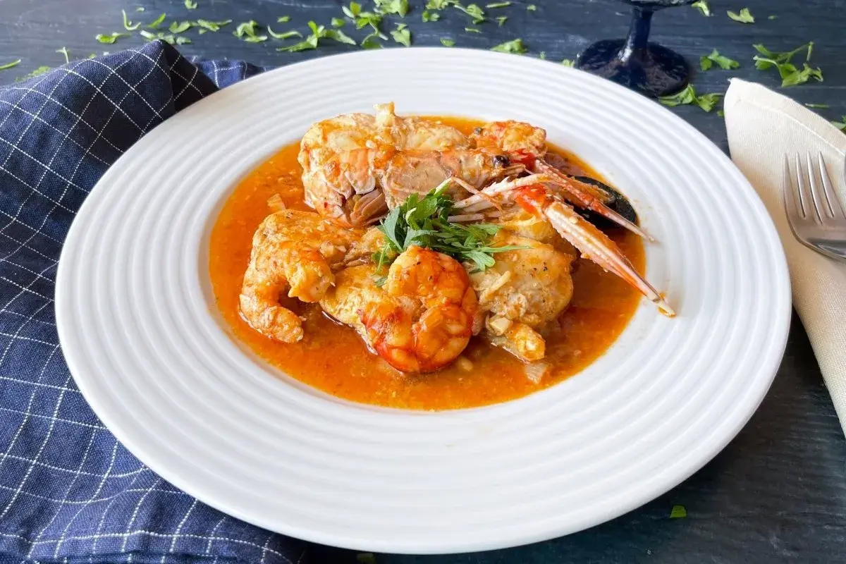 Un plato de rap a la marinera con gambas y cigalas en una salsa rojiza, decorado con perejil fresco.