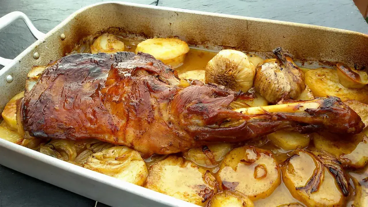 Paletilla de cordero asada con patatas y ajos dorados en una bandeja. Jugosa y apetitosa, lista para servir.