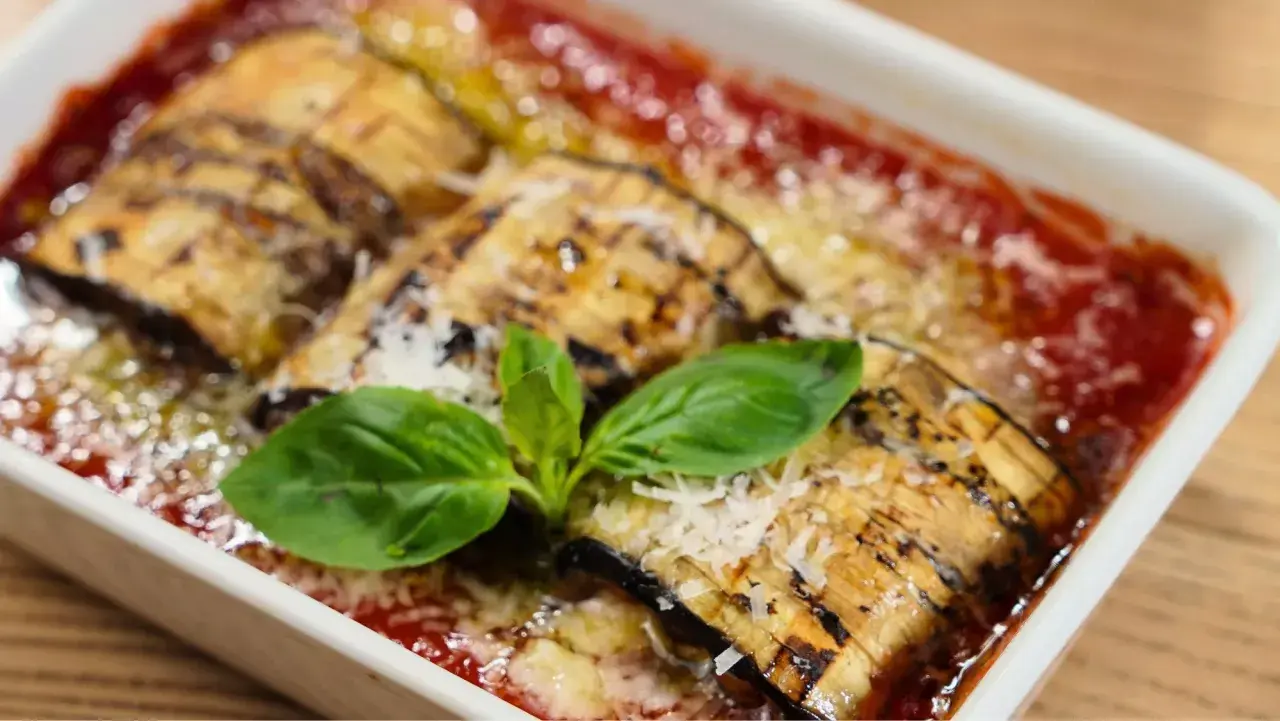 Delicioso pastel de berenjenas gratinado con queso y bañado en salsa de tomate, adornado con hojas de albahaca fresca.