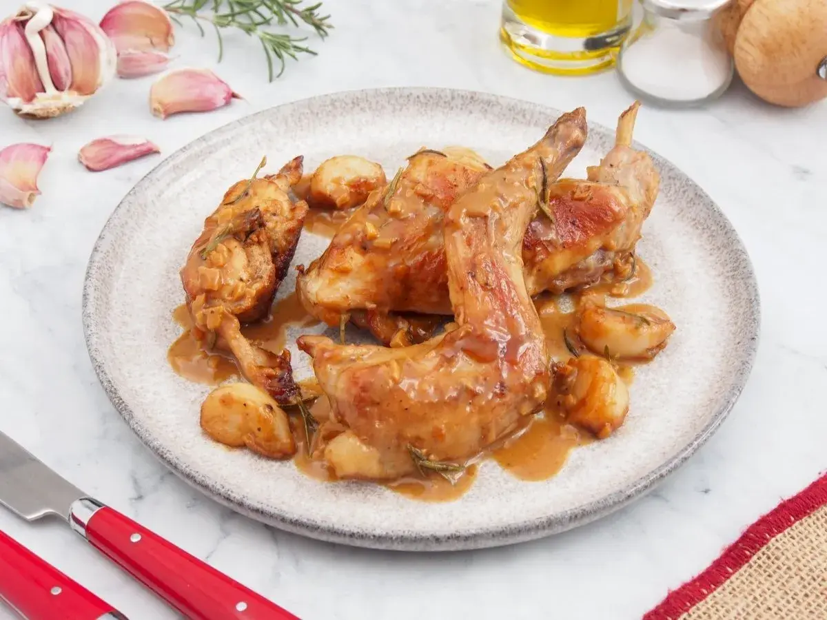 Delicioso conejo al ajillo servido en un plato, con ajos enteros asados y salsa dorada.