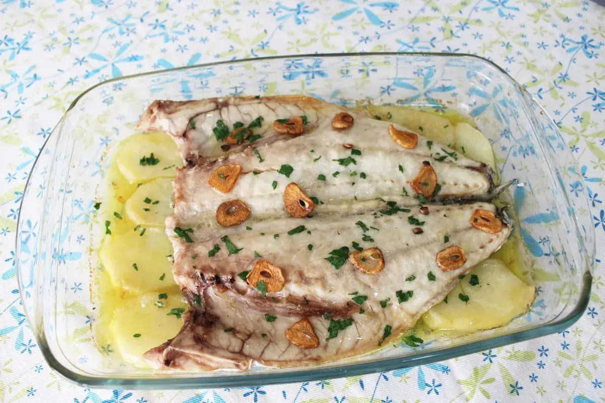 Lubina a la espalda horneada con patatas, ajo y perejil en una fuente de cristal.