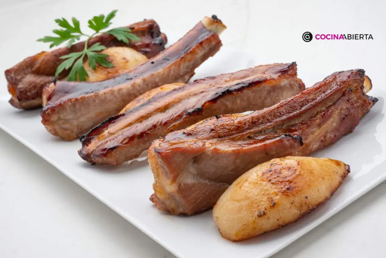 Costillas de cerdo jugosas y doradas, servidas en una bandeja blanca con patatas asadas y una ramita de perejil.