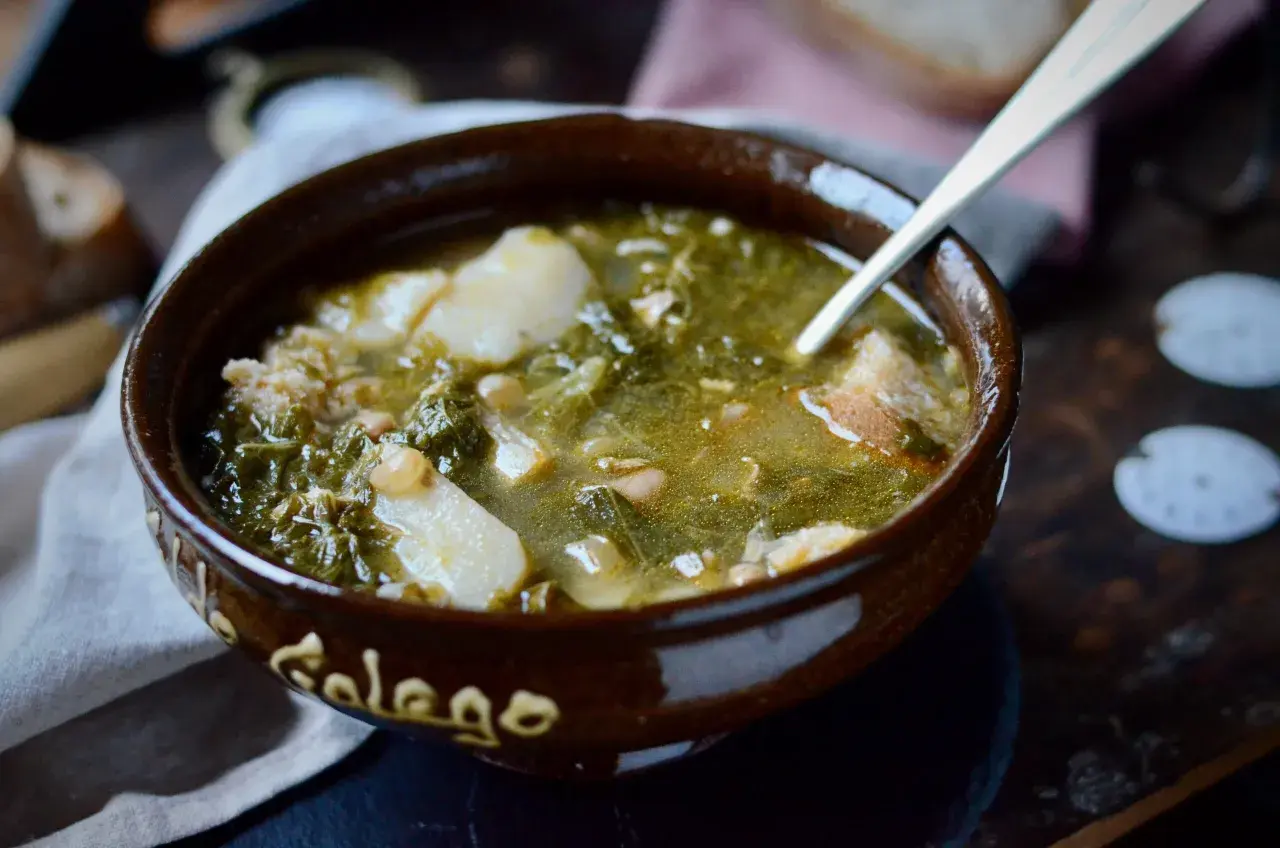 Un cuenco de barro con la palabra "Gallego" escrita, lleno de un reconfortante caldo gallego con patatas, grelos y trozos de pan.